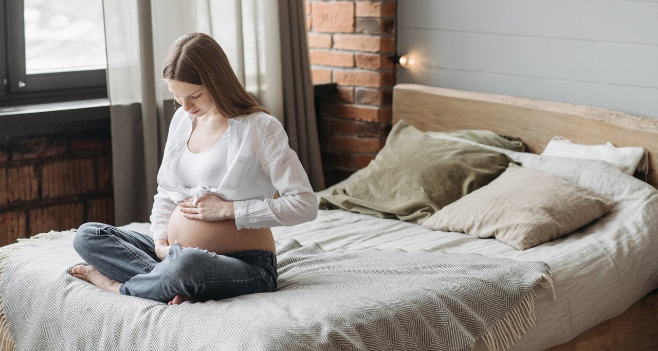 Mulher grávida sentada na cama olhando sua barriga.