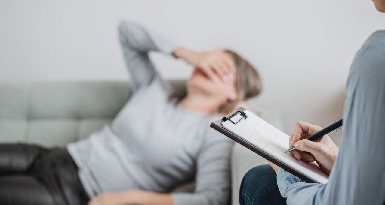 Paciente deitada em um sofá, enquanto seu psiquiatra faz anotações na prancheta durante um diagnóstico de depressão.