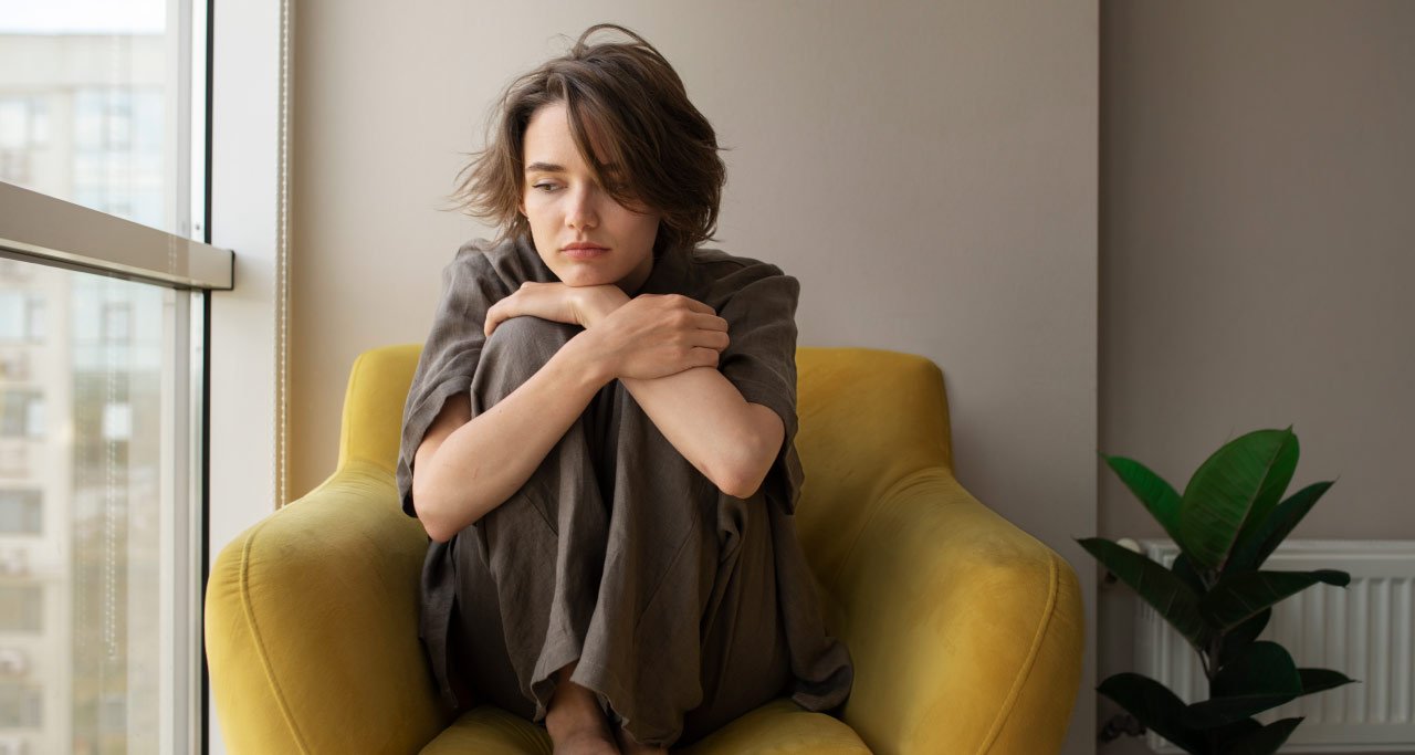 Mulher com depressão sentada no sofá, ao lado da janela, abraçando os próprios joelhos.