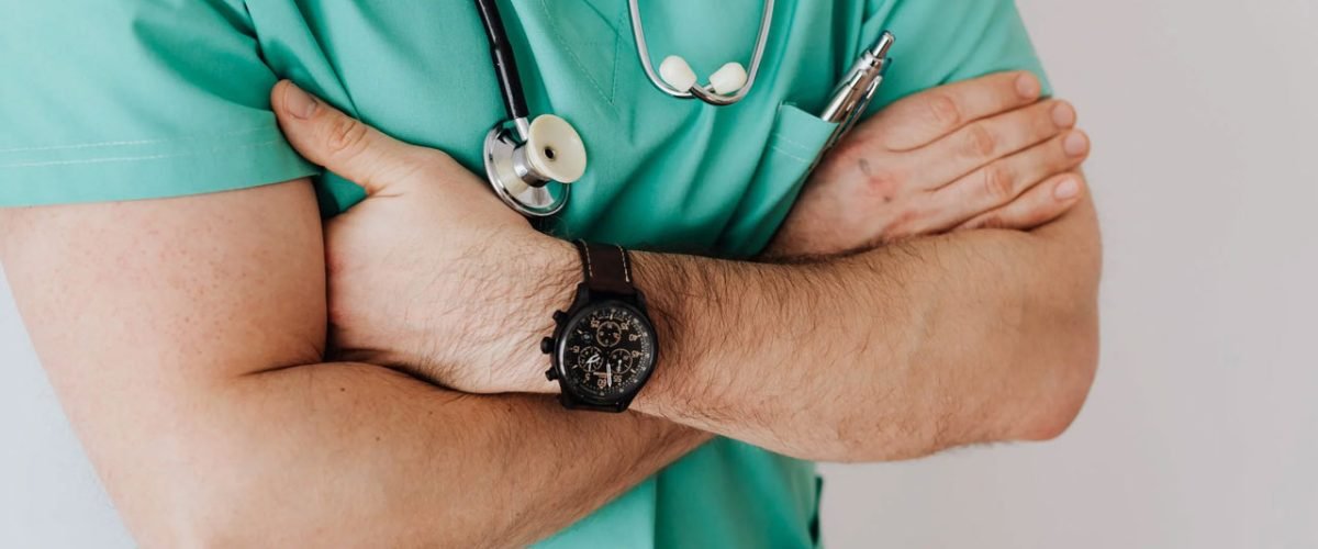 Médico vestido de jaleco verde, de braços cruzados e com estetoscópio entre o pescoço.