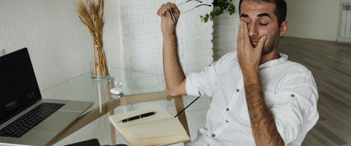 Homem em mesa de trabalho aparentemente cansado, passando a mão nos olhos.