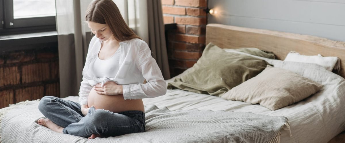 Mulher grávida sentada na cama olhando sua barriga.